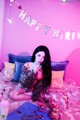 A woman sitting on a bed with balloons and a birthday hat.